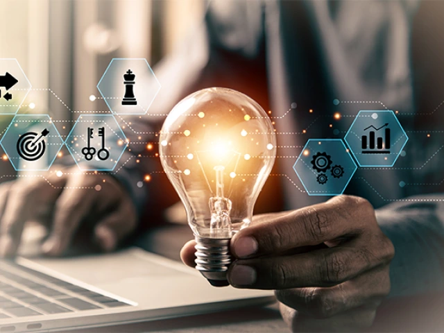 AI-generated image of a person holding a lightbulb with one hand, and typing on a laptop with the other. Image is overlaid with small octagon-shaped icons symbolizing the generation of ideas.